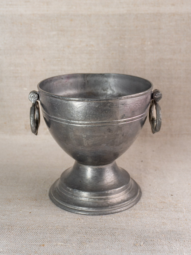 Antique French pewter footed bowl with ornate handles, wide basin, on textured beige fabric background.
