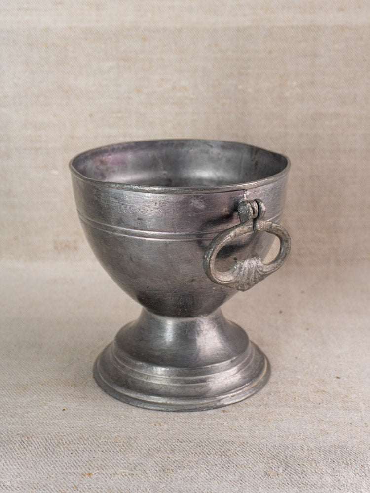 Antique French pewter footed bowl with pedestal base and side metal ring, shown on neutral textured surface.