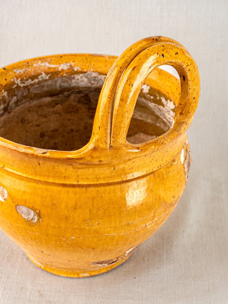 19th-century French yellow-glazed terracotta hanging planter with tall curved handle and chips, on neutral fabric background.