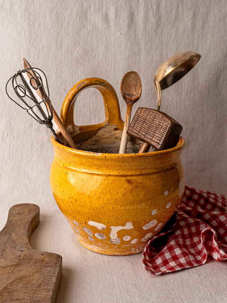 Antique French yellow glazed terracotta hanging planter with vintage kitchen utensils, beside wooden board and red checkered cloth.