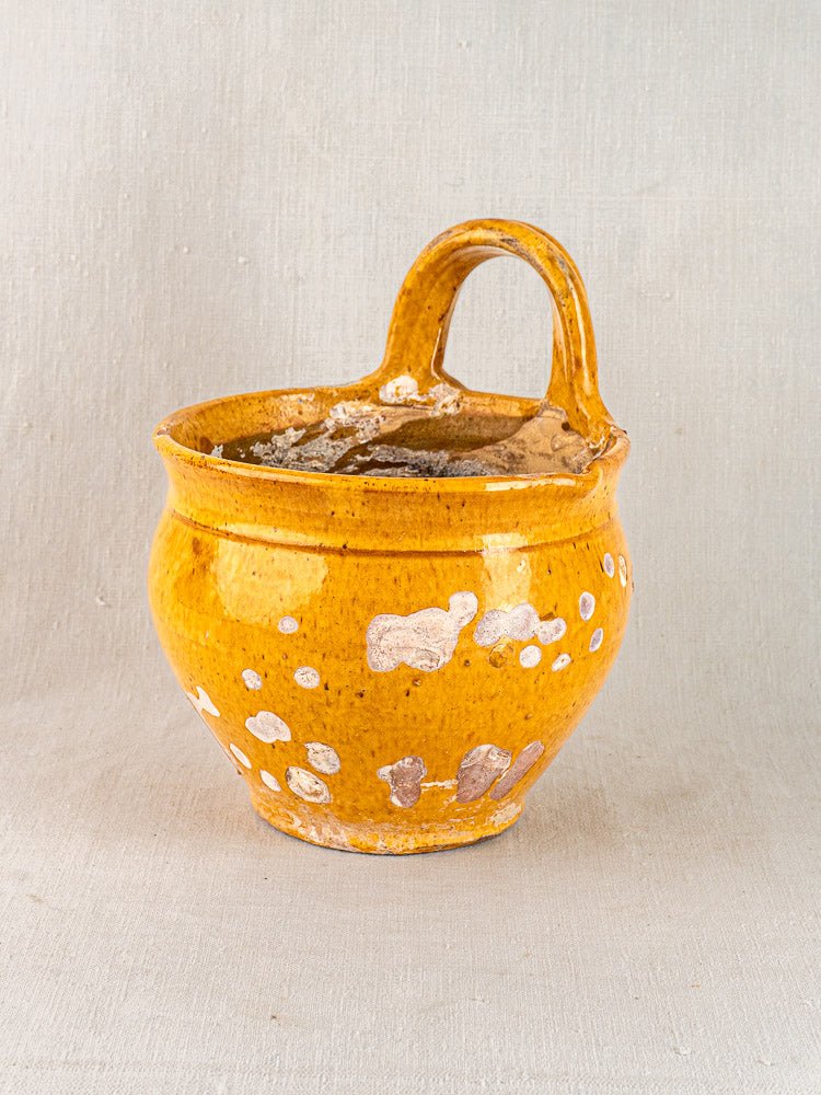 Antique French yellow glazed terracotta pichet with white spots and handle, rustic wear, on neutral fabric background.