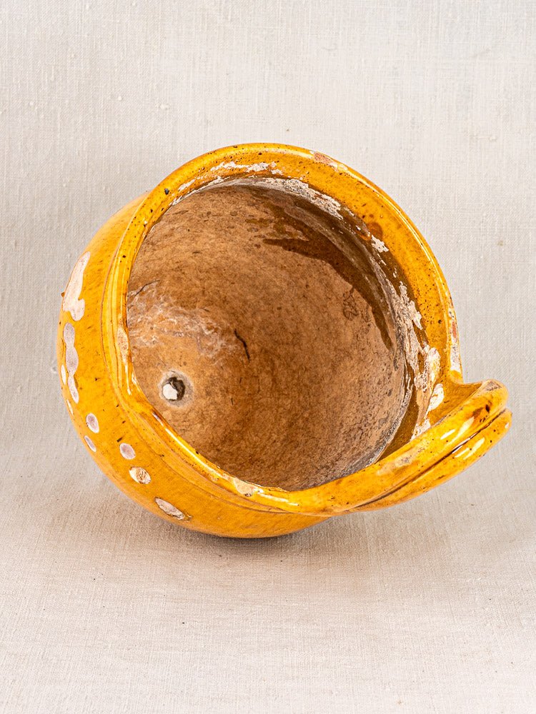 Antique French yellow-glazed terracotta hanging planter with spout and handle, rustic patina, on neutral fabric.