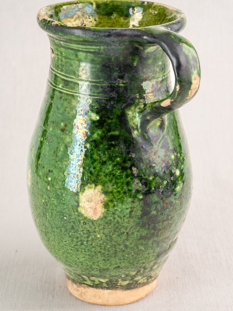 19th-century French green-glazed terracotta pitcher with handle, worn glaze, exposed clay, neutral background.