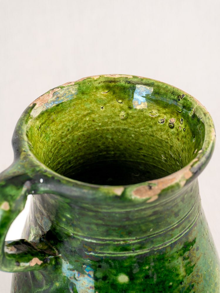 Antique French green glazed terracotta pitcher with chipped edges, glossy finish, and interior water droplets.