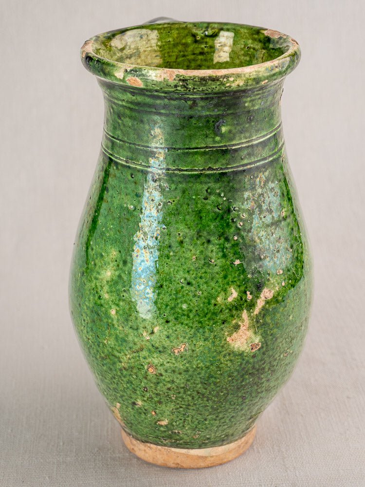 19th-century French green glazed terracotta pitcher with weathered rim and base on neutral background.