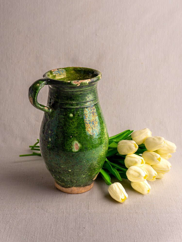 Antique French green glazed terracotta pitcher with handle beside white tulips on fabric background.