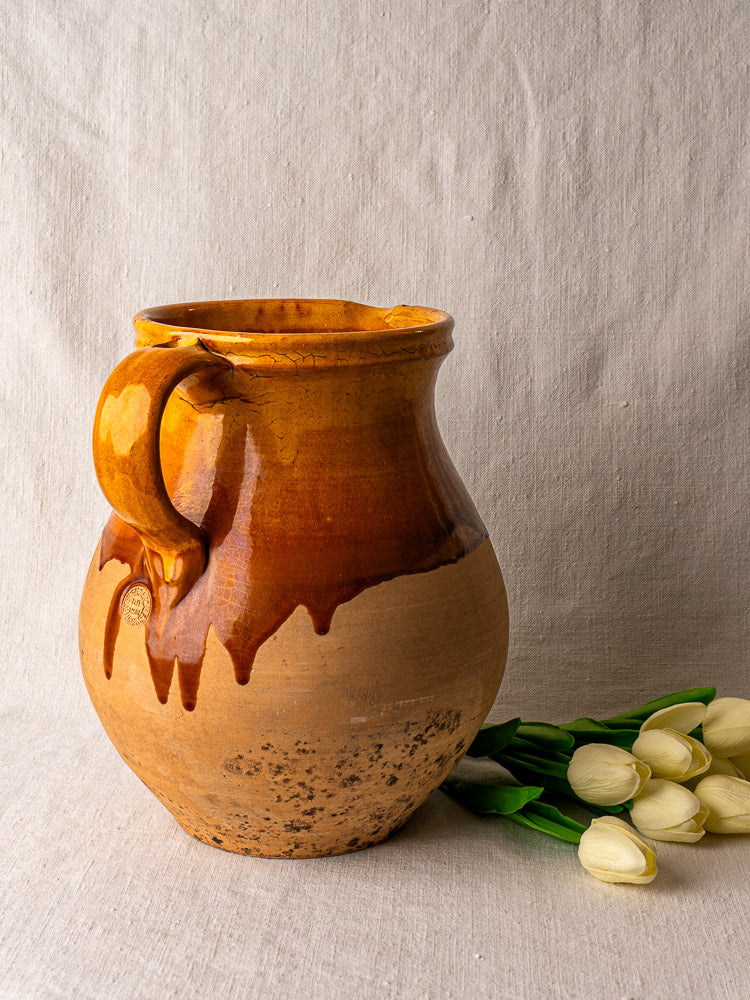 French Provençal terracotta pitcher with single handle, amber-glazed top, on neutral fabric background.