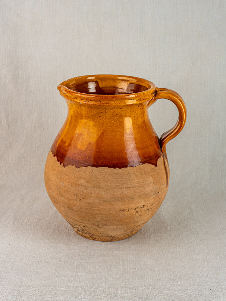 French Provençal terracotta pitcher with single handle, amber-glazed top, on neutral fabric background.