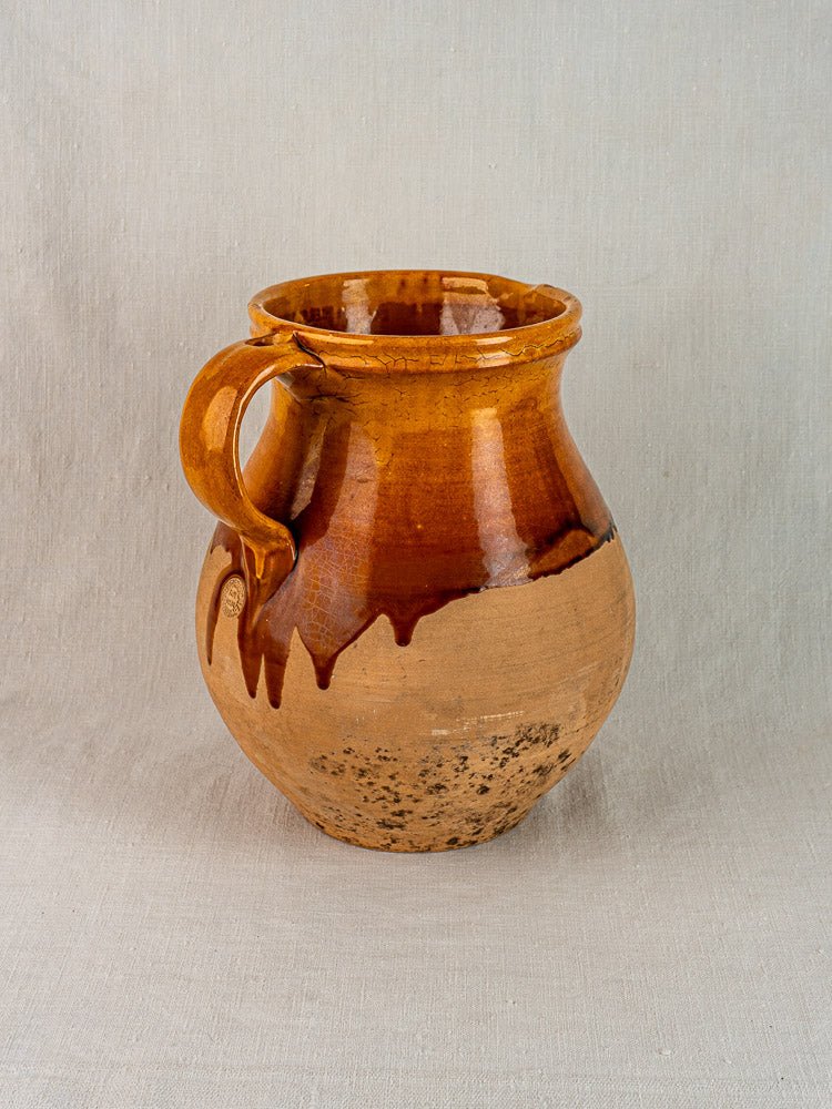 French Provençal terracotta pitcher with single handle, amber glaze, on light fabric surface.