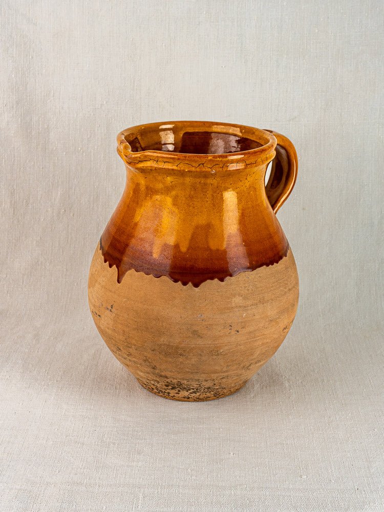 French Provençal terracotta pitcher with amber-glazed upper, single handle, on neutral fabric background.