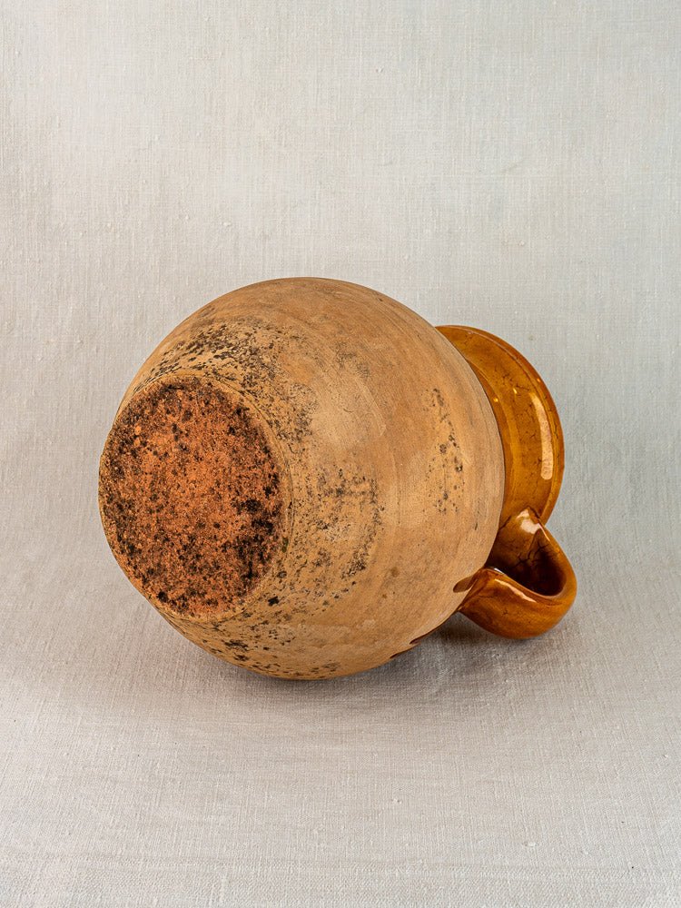 French Provençal terracotta pitcher with amber glaze, rustic round base, and visible wear on fabric background.
