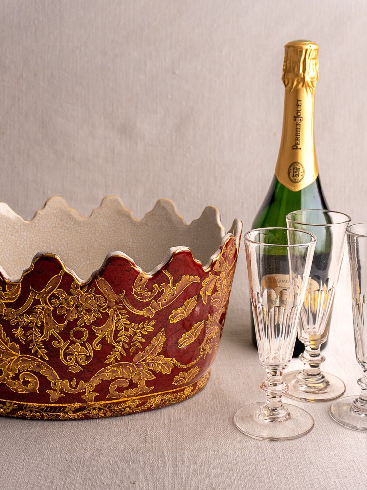 Antique French Terre de Fer wine cooler with red and gold accents beside champagne on neutral fabric.