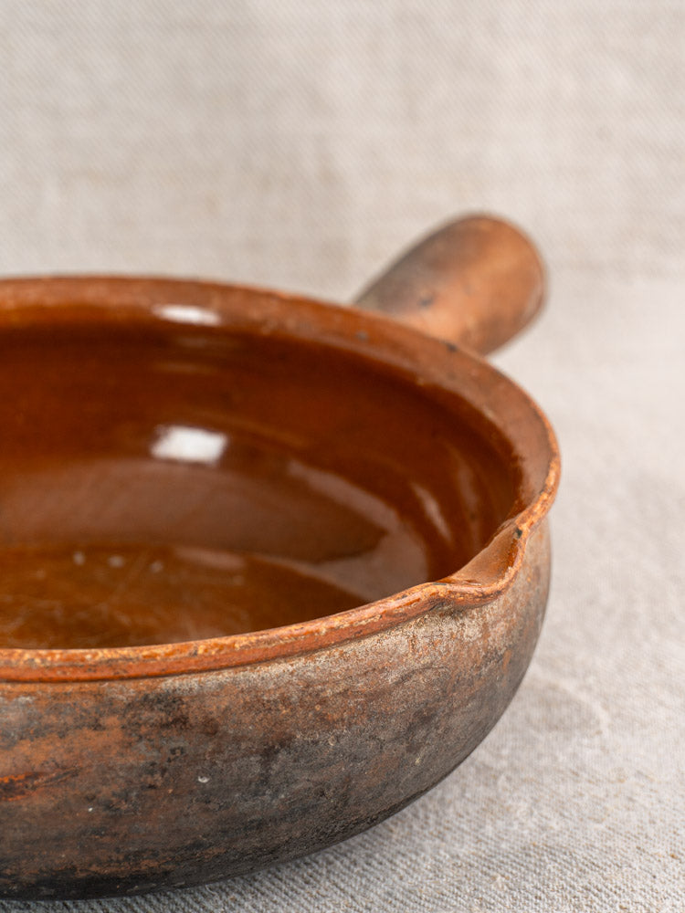 Antique French Saint-Uze marked stoneware poêlon, rustic brown ceramic pan with handle and spout on beige fabric.