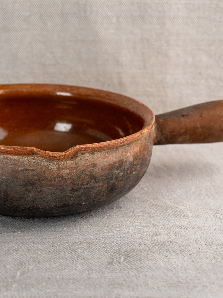 Antique French Saint-Uze stoneware poêlon with brown glaze, side pour spout, and elongated handle on beige fabric background.