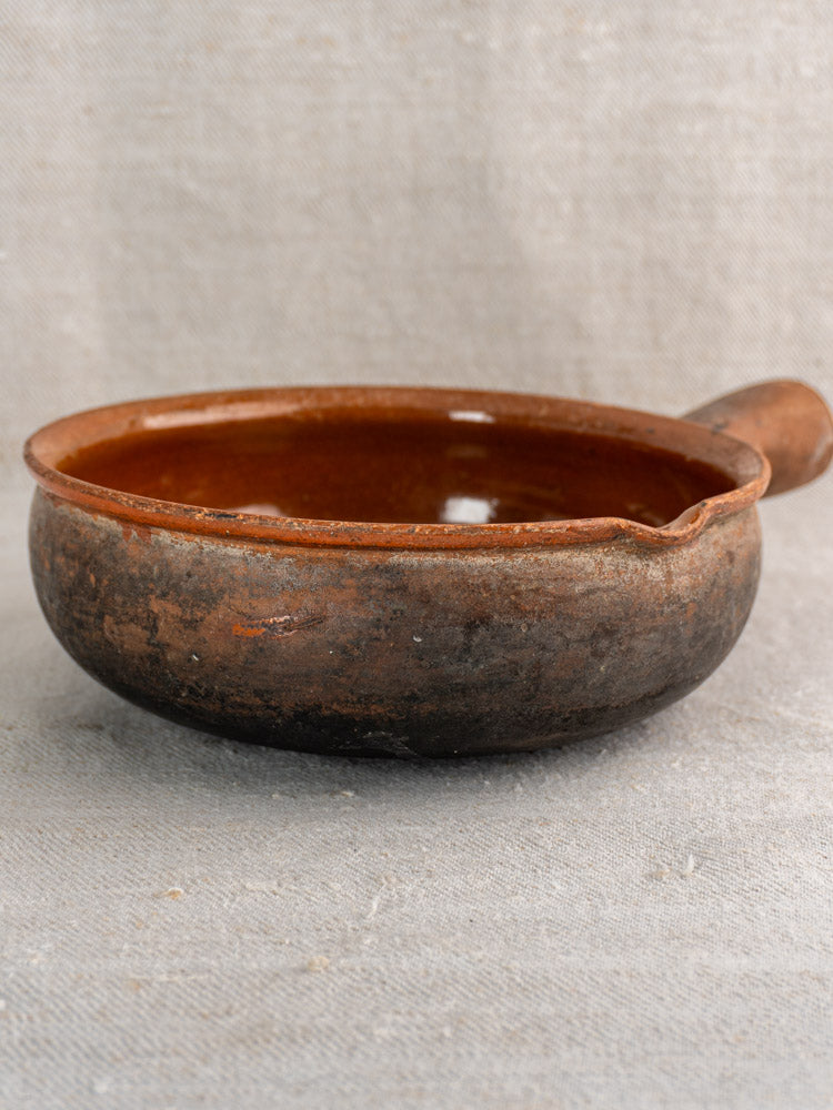 Antique French Saint-Uze stoneware poêlon with handle, worn outside, glossy inside, on neutral fabric.