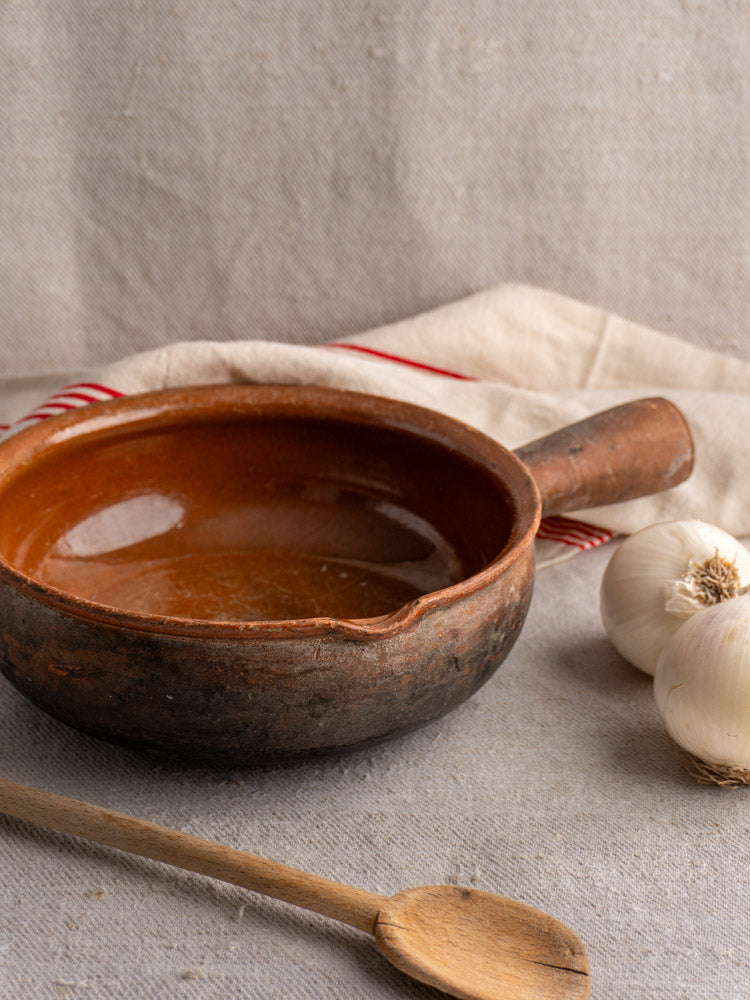 Antique French Saint-Uze marked stoneware poêlon with rustic clay handle, on beige cloth with garlic and wooden spoon.