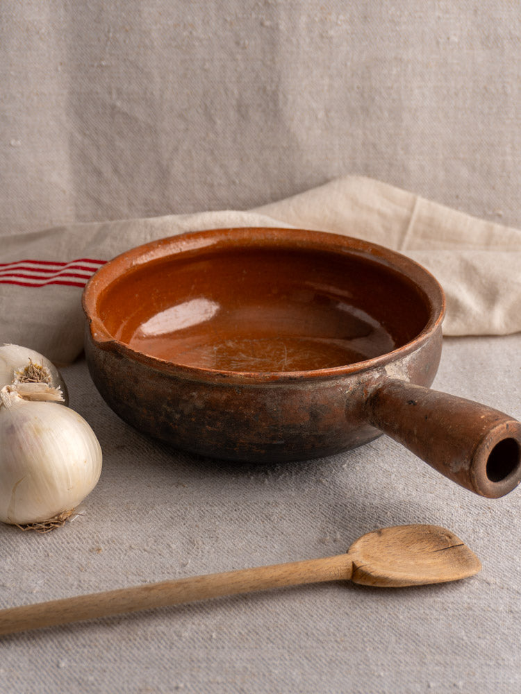 Antique French Saint-Uze marked stoneware poêlon with long handle, shown beside garlic and wooden spoon.