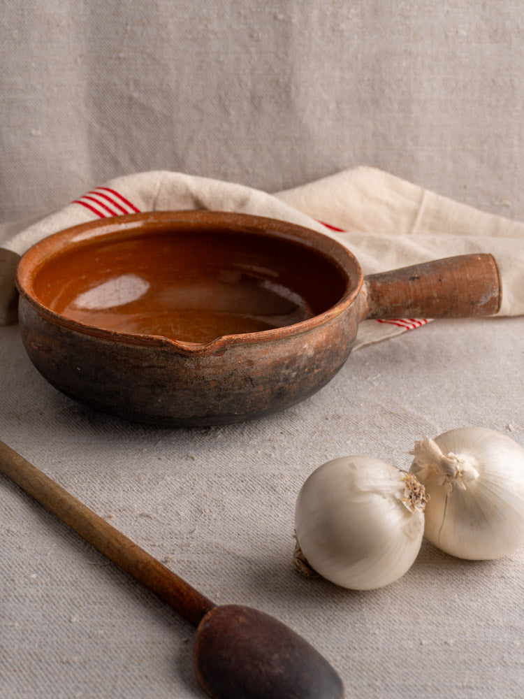 Antique French Saint-Uze stoneware poêlon cooking pan with garlic bulbs, wooden spoon, and striped cloth.