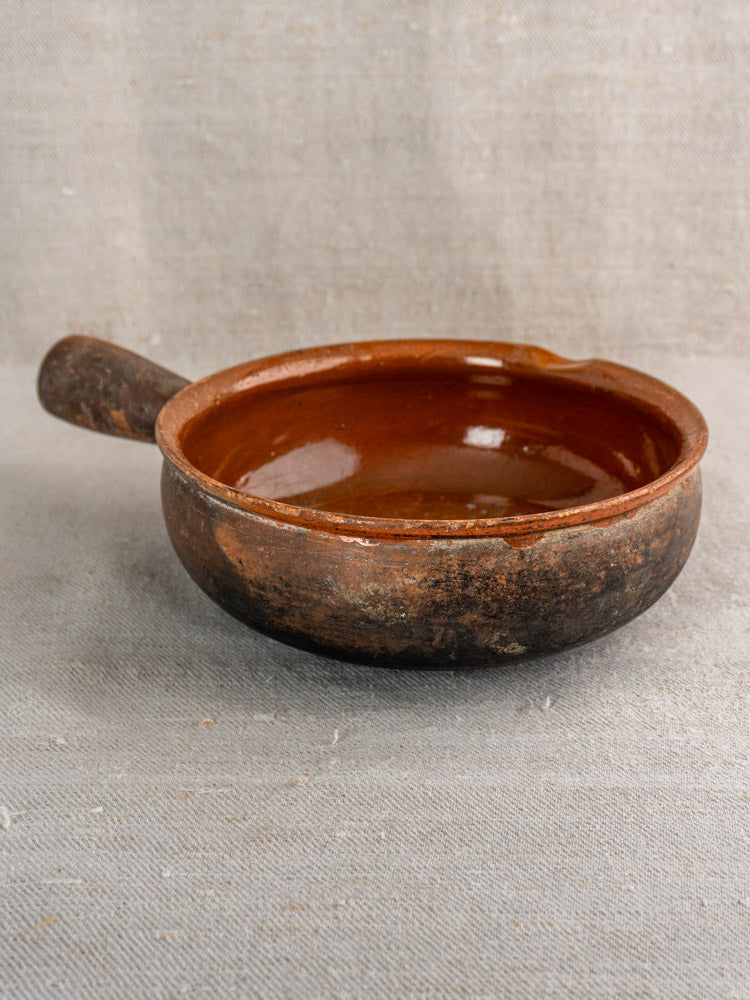 Antique French Saint-Uze stoneware poêlon with handle, rustic brown exterior, glazed interior on light fabric.
