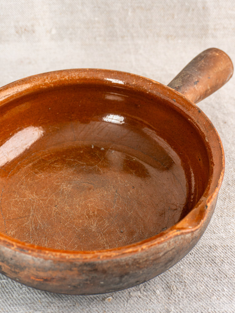 Antique French Saint-Uze stoneware poêlon with handle and aged patina on beige textured fabric.