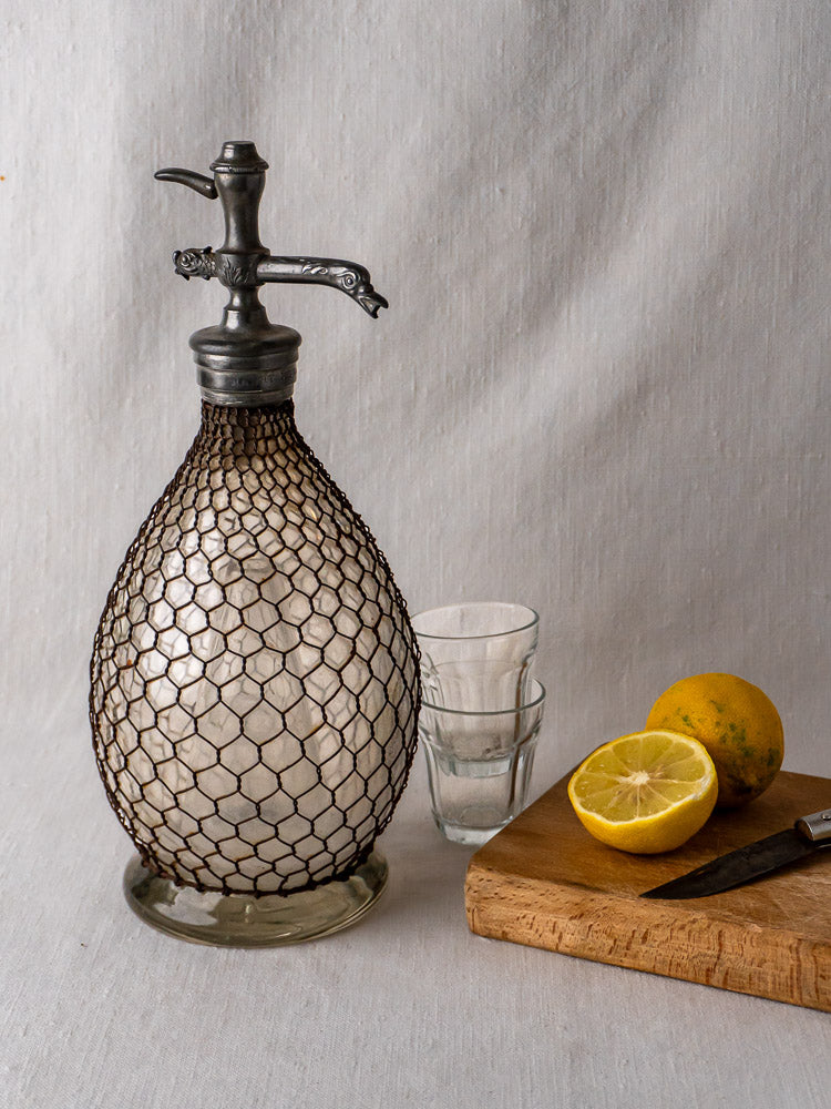 Antique French Henry Paris seltzer siphon with wire mesh, paired with glasses and lemons on board.