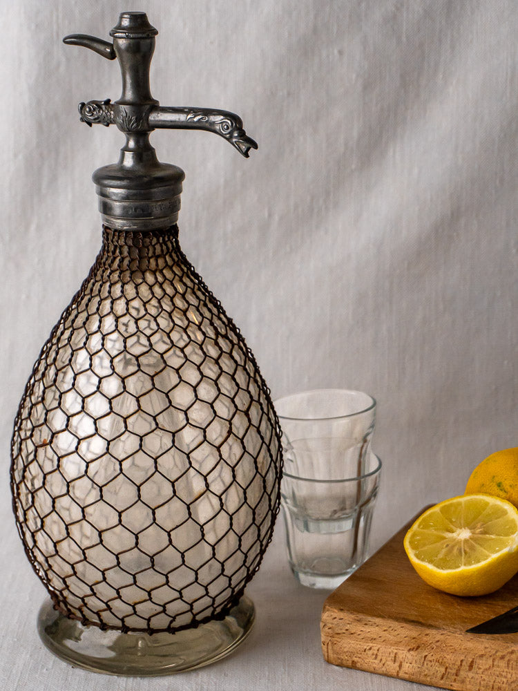 Antique French wire mesh seltzer siphon with two glasses and lemons on wood board.