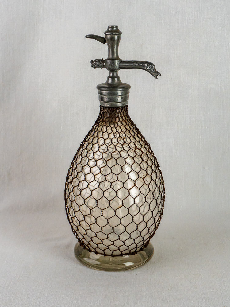 Antique French Henry Paris seltzer siphon with wire mesh and metal spout on neutral fabric.