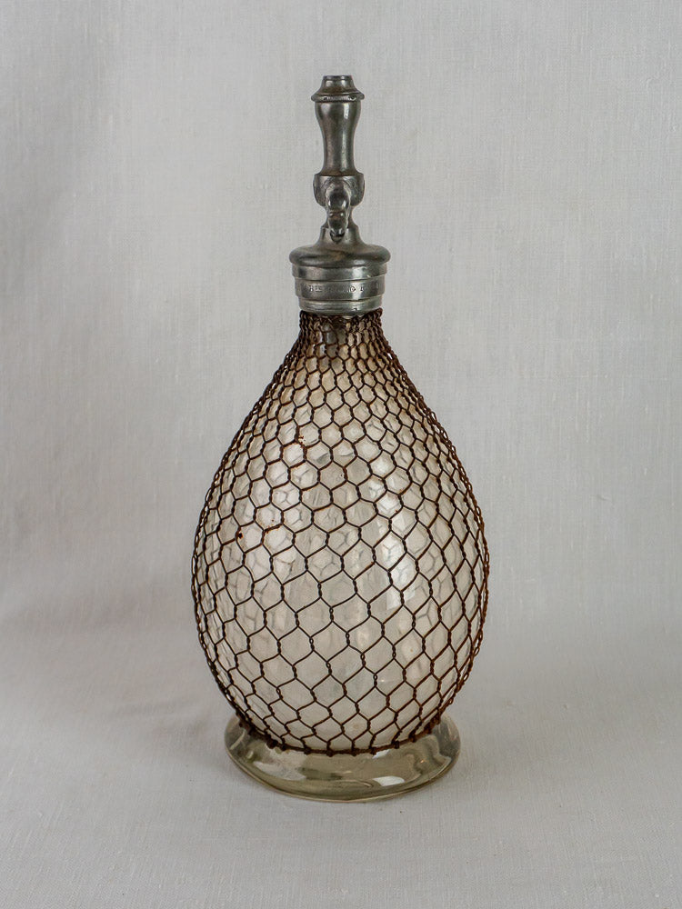 Antique French glass seltzer siphon with metal stopper, wrapped in wire mesh, on a light background.