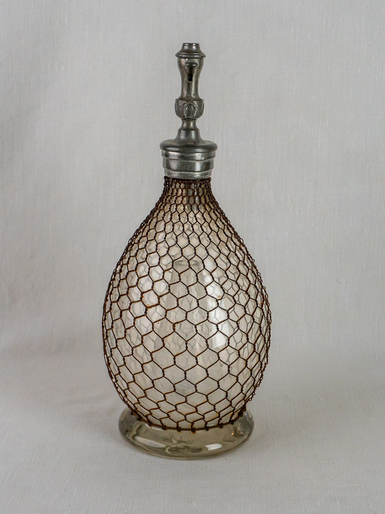 Antique French Henry Paris seltzer siphon with wire mesh, metal stopper, and rounded clear glass body.