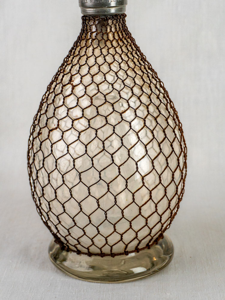 Antique French glass seltzer siphon with brown wire mesh, displayed on a white surface.