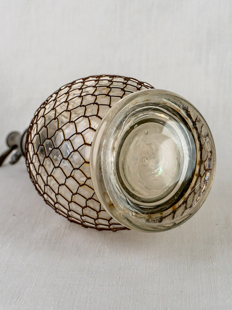 Antique French glass seltzer siphon with brown wire mesh, displayed on white background.