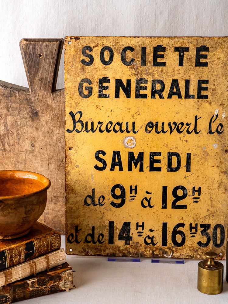 Antique French Société Générale Bank hand-painted steel sign with black text on yellow, rustic wood backdrop. Old books, brown bowl, brass object in foreground.