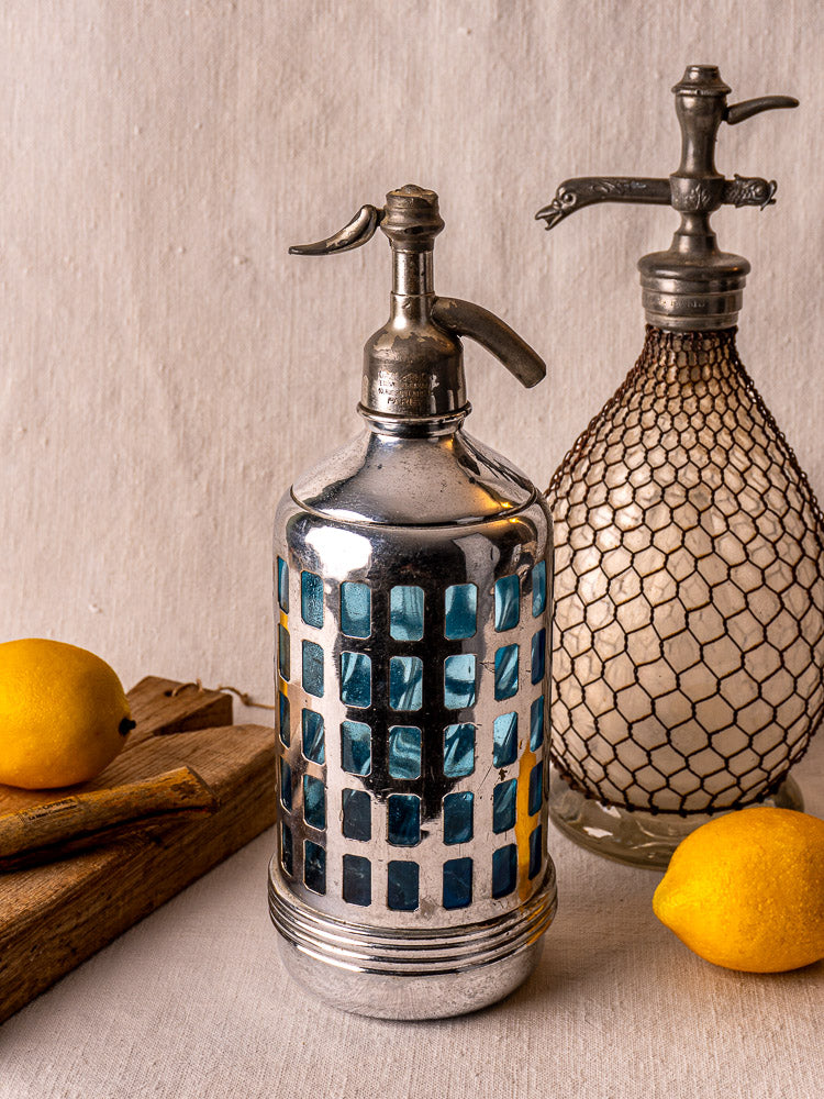 Antique French Gazo-Siphon soda siphon with blue-silver mesh detailing beside lemons on cloth-draped table.