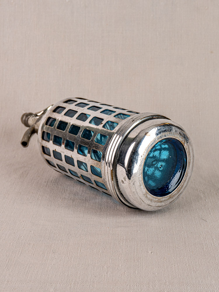 Antique French blue glass soda siphon with silver metal grid, Paris bistro style, on neutral fabric background.