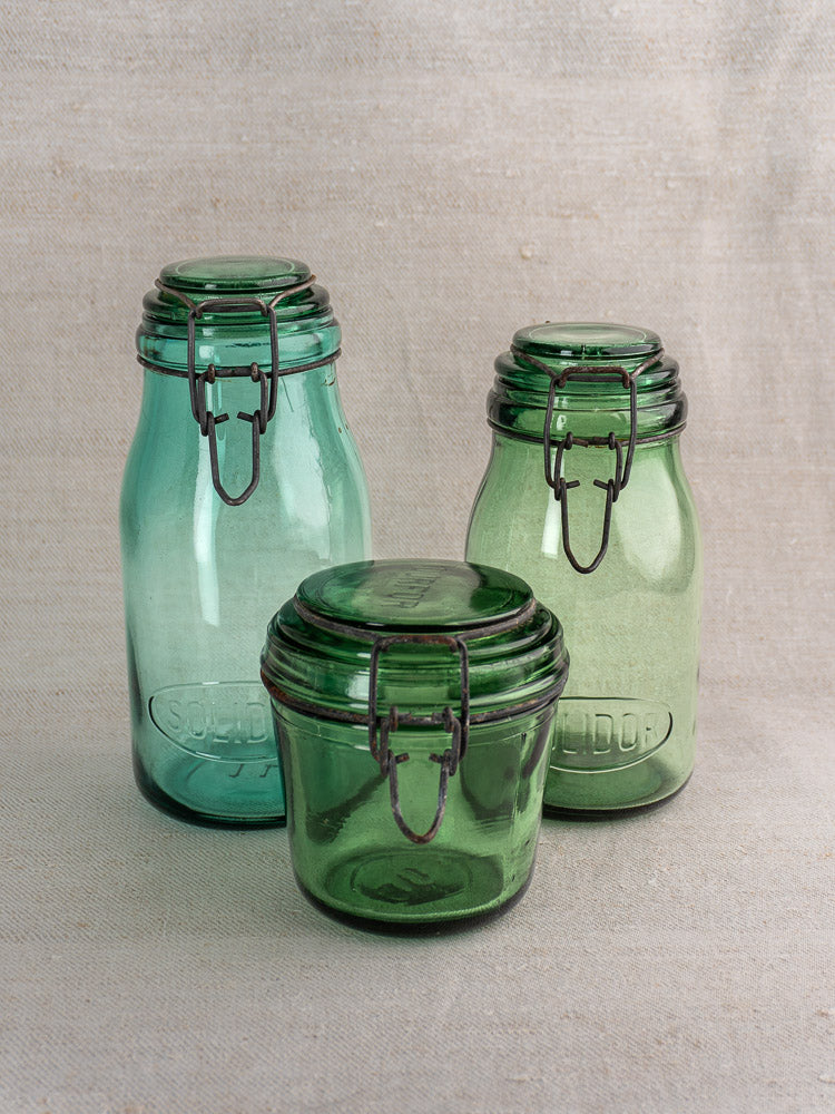 Set of three antique French Solidor & Durfor preserve jars with embossed fronts and metal clasps on neutral fabric.