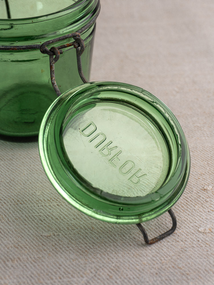Antique French green glass preserve jar with embossed lid and metal clasps on textured fabric.