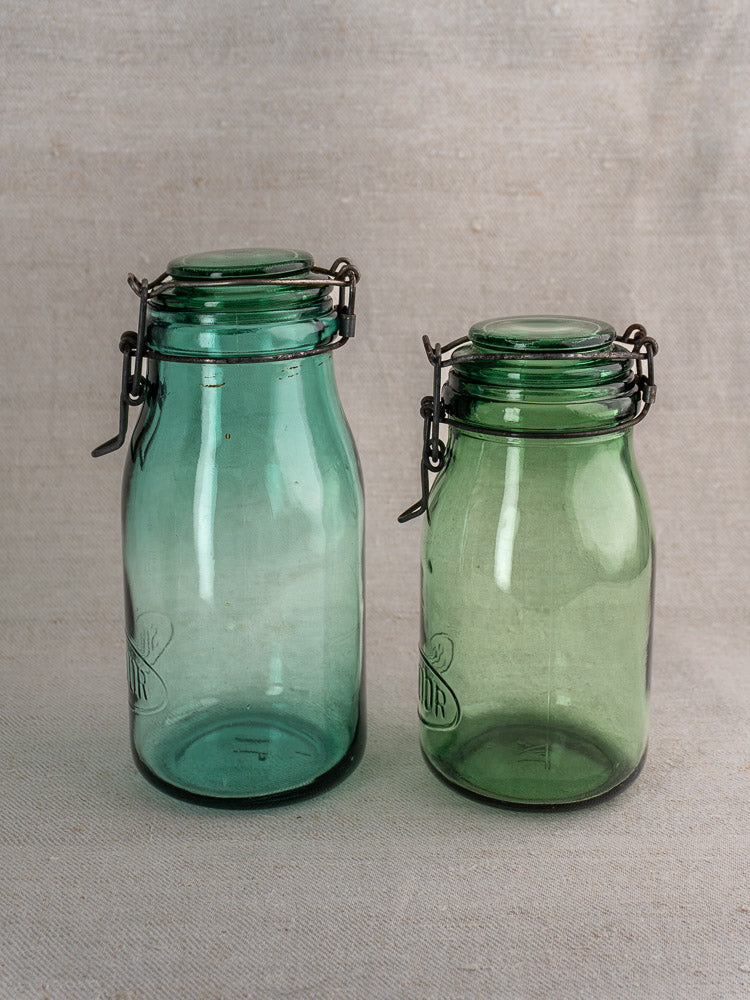 Set of three antique French Solidor & Durfor greenish glass preserve jars with metal latch lids on beige fabric.