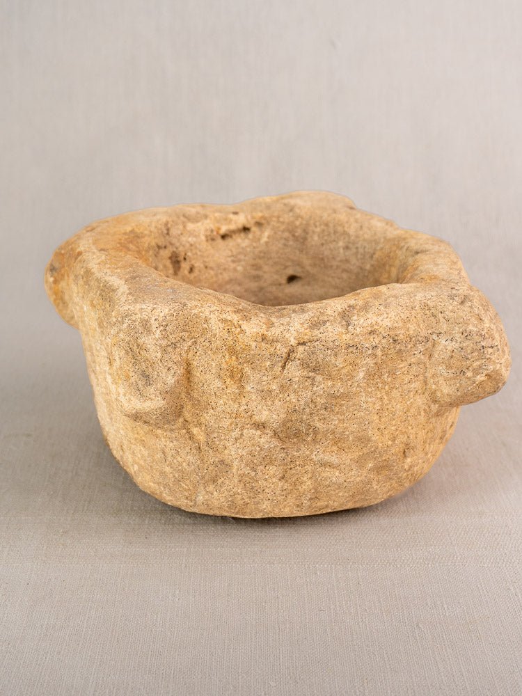 Antique French stone mortar and pestle with irregular edges and textured beige surface on neutral fabric background.