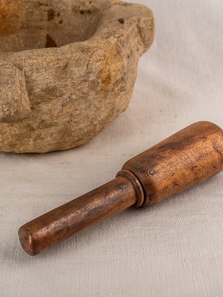 Antique French stone mortar and pestle with weathered wood, hand-carved details, on a light fabric surface.