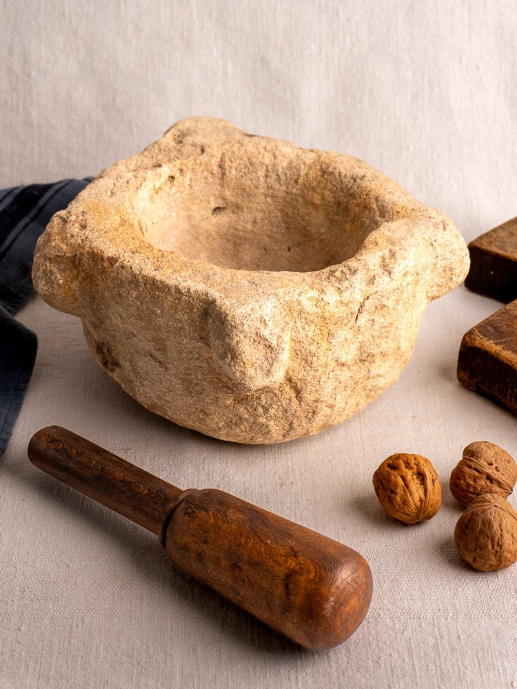 Antique French stone mortar and wooden pestle with walnuts on light fabric, wooden boards in background.