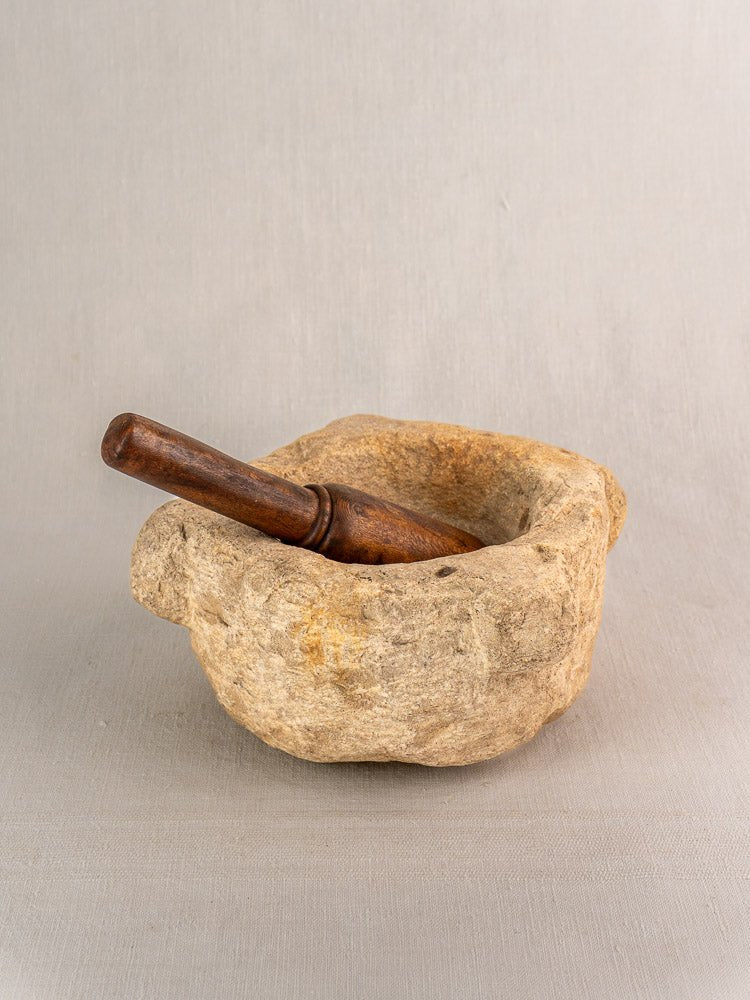 Antique French stone mortar with wooden pestle, displayed on a light gray textured surface, neutral backdrop.