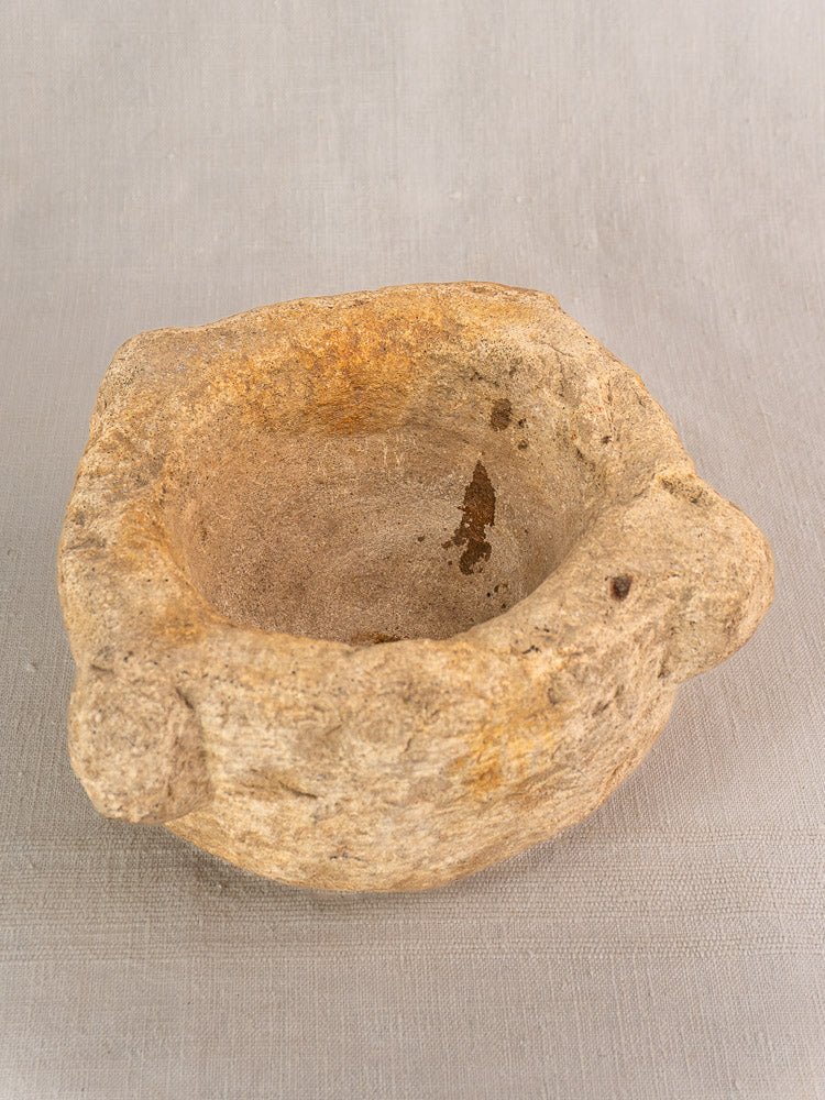 19th-century French stone mortar and pestle with uneven edges and small handles on light gray textured surface.