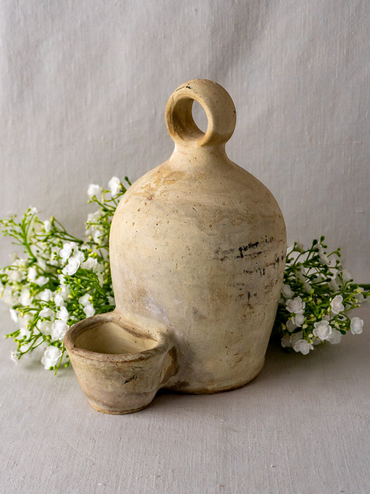 Rustic French stoneware bird waterer with round handle and cup, set against white flowers.