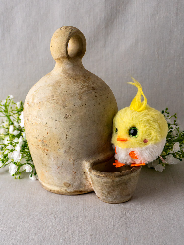 Antique French stoneware bird waterer with plush yellow chick, rustic ceramic jug, and cup on light fabric.