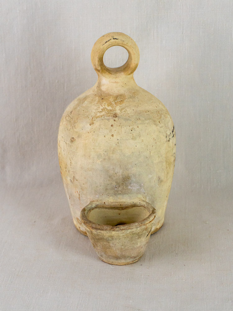 Antique French stoneware bird waterer with round handle and matching cup on neutral fabric background.