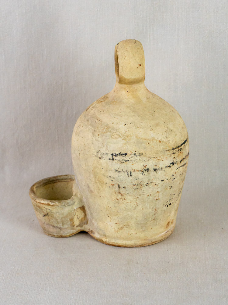 Antique French stoneware bird waterer with looped handle and base bowl, on light fabric background.