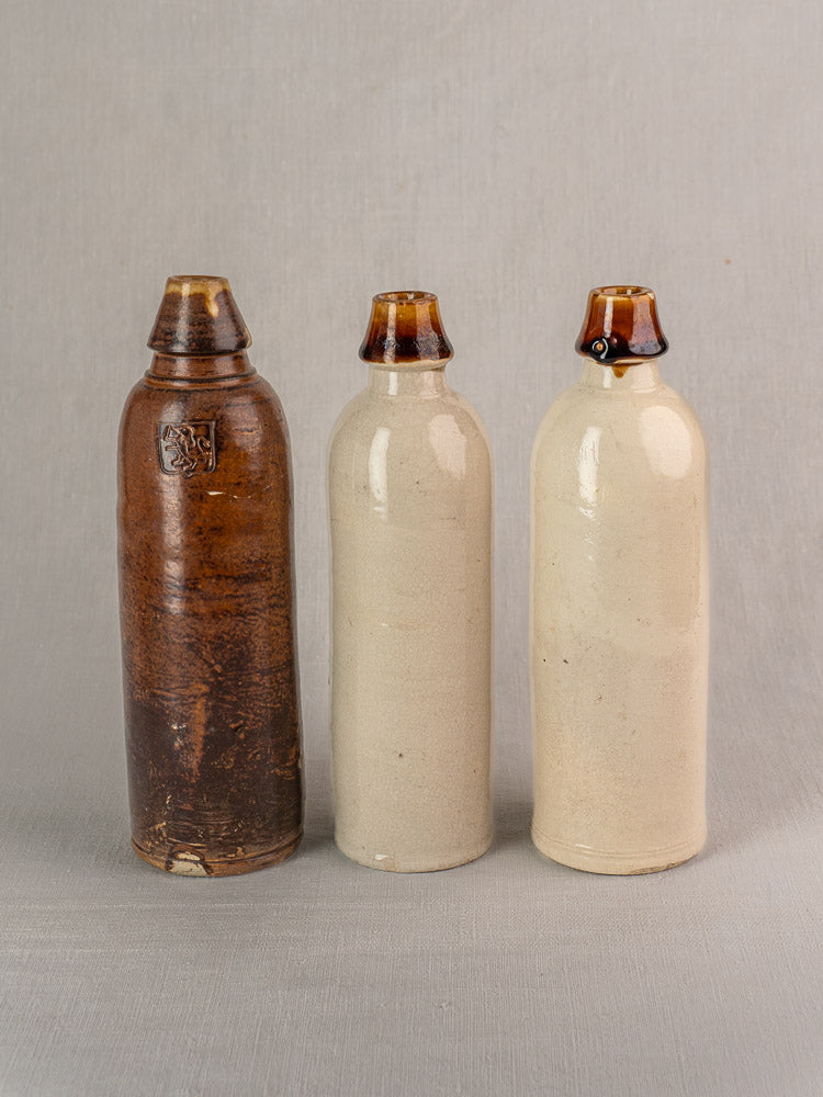 Set of three antique French stoneware cider and beer bottles with contrasting glazed finishes on gray fabric.