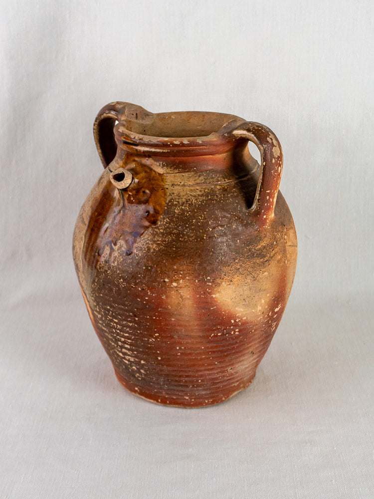 Antique French stoneware oil jug with ochre glaze, two handles, and spout on fabric background.