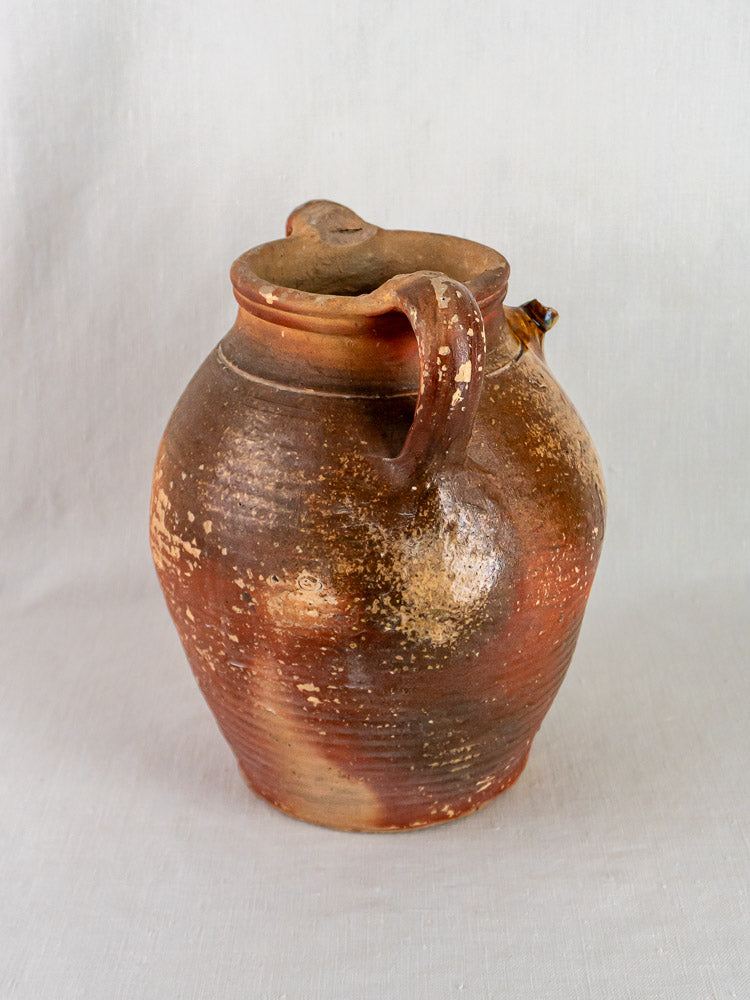 Antique French stoneware oil jug with ochre glaze, handle, spout, and textured surface on light background.