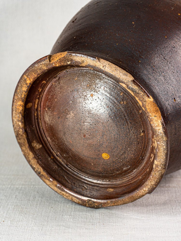 Antique French pot à graisse in salt-glazed stoneware with rustic dark brown surface and rim wear, on woven fabric.
